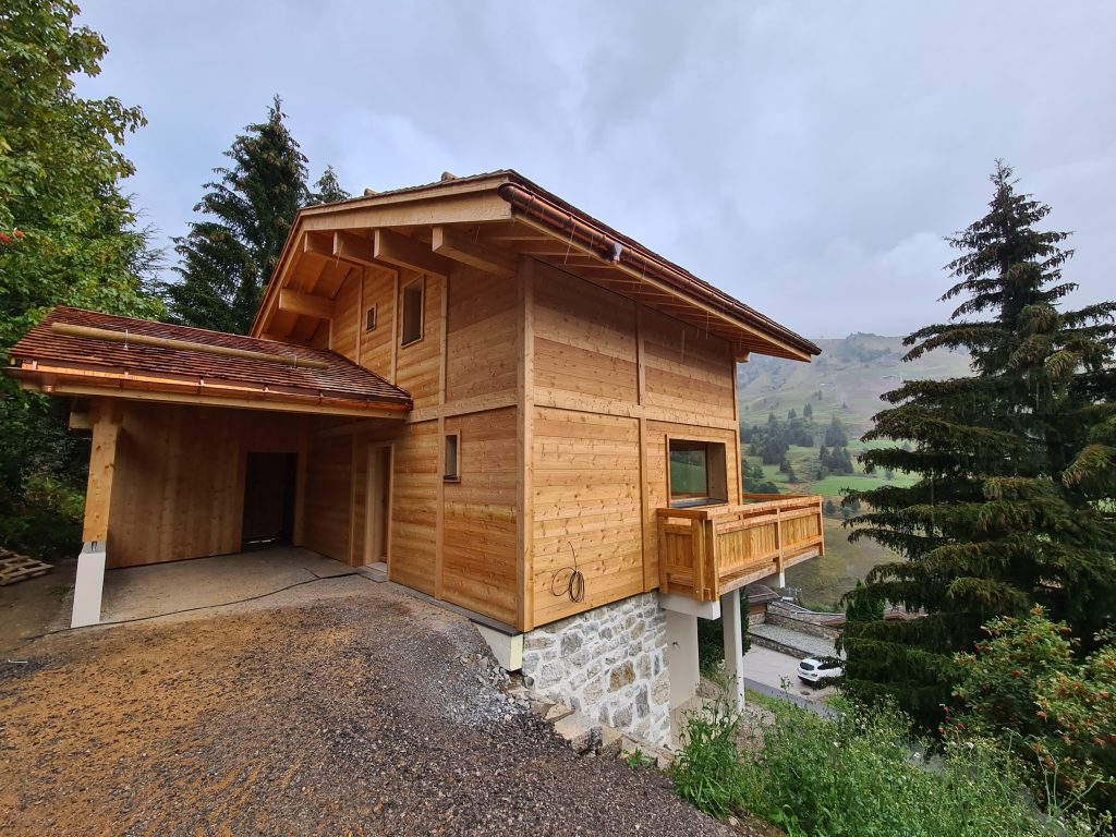 Maison à ossature bois située à Thônes, en Haute-Savoie, réalisée par DDLP Structures Bois. Charpente traditionnelle avec toiture en bois et balcon en bois massif. Vue sur la montagne environnante, mettant en valeur l'utilisation de matériaux locaux et durables pour une construction éco-responsable.
